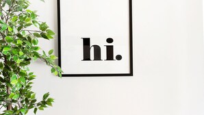 Ein gerahmtes Schild mit 'hi', eine Zimmerpflanze, eine Bank und zwei gewebte Körbe an einer weißen Wand.