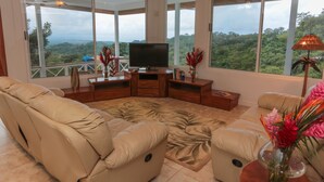 A spacious living room with a large flat-screen TV, beige sofas, a patterned rug, and a view of lush greenery through large windows. - Casa Papillon: Waterfall pool, Jungle Views, Terrace, Sleeps 12 (Manuel Antonio)