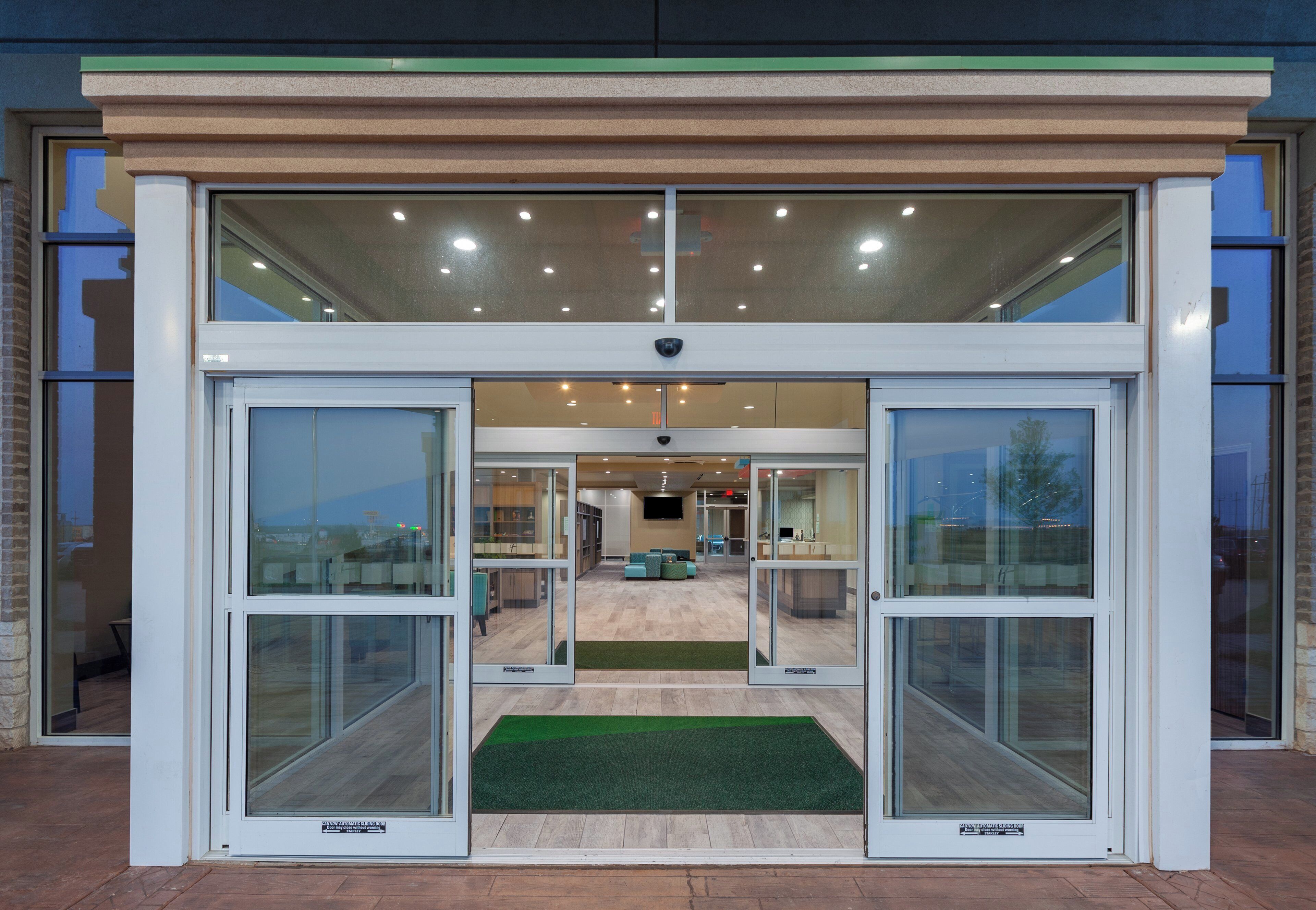 Entrada de un edificio moderno con puertas correderas automáticas, un felpudo verde y vista al interior que incluye zonas de estar y un mostrador de recepción.