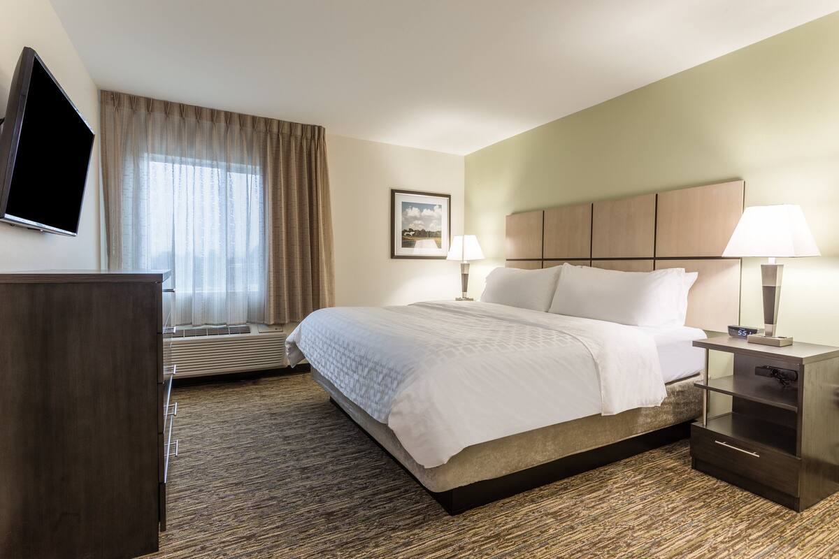 Une chambre d’hôtel avec un lit, des lampes de chevet, un téléviseur à écran plat, une fenêtre avec des rideaux et un tableau encadré au mur.