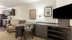 A hotel room with a kitchenette, a dining area with a table and chairs, a flat-screen TV, a lamp, and a framed picture on the wall.