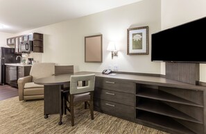 A hotel room with a kitchenette, a dining area with a table and chairs, a flat-screen TV, a lamp, and a framed picture on the wall.