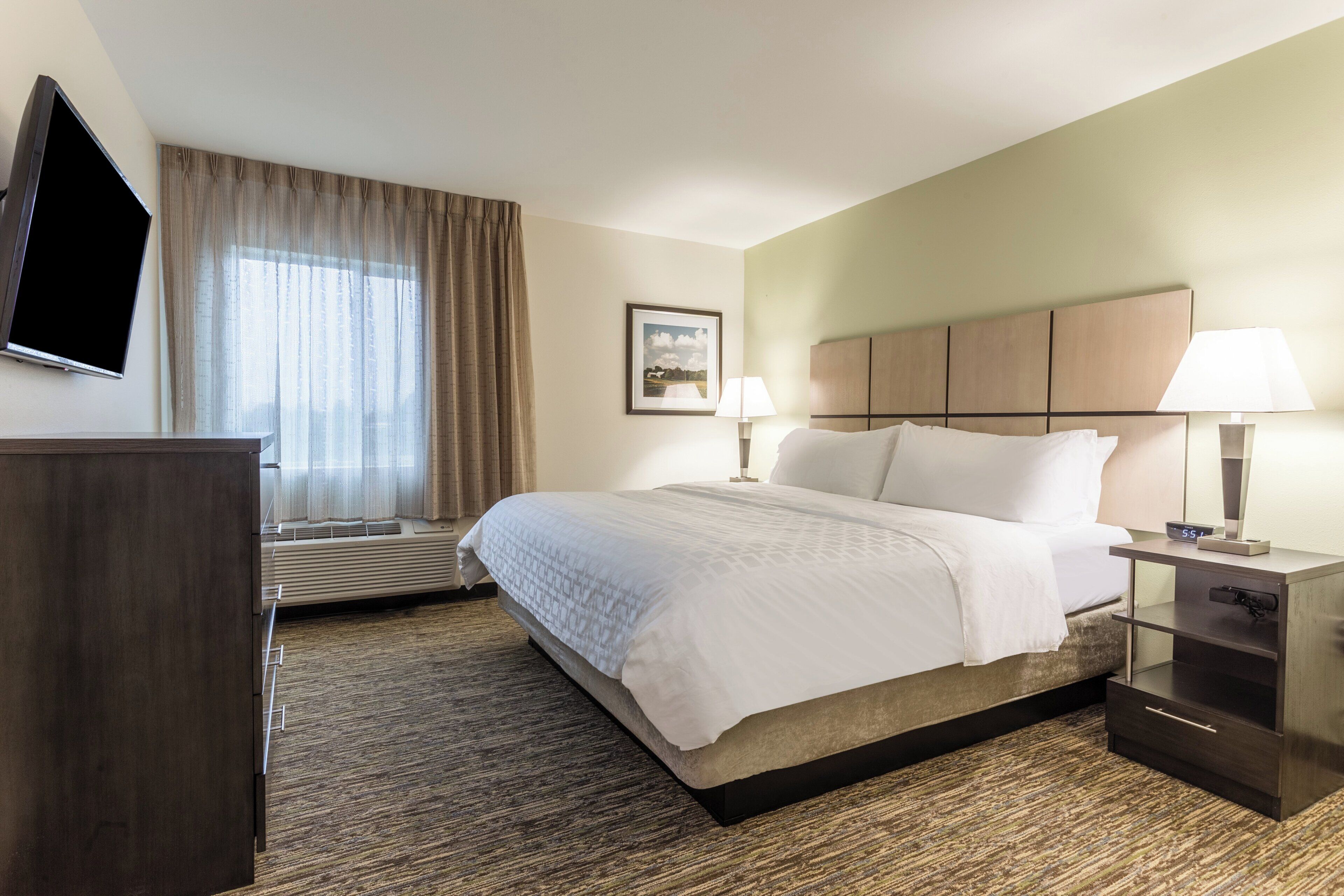 A hotel room with a bed, bedside lamps, a flat-screen TV, a window with curtains, and a framed picture on the wall.