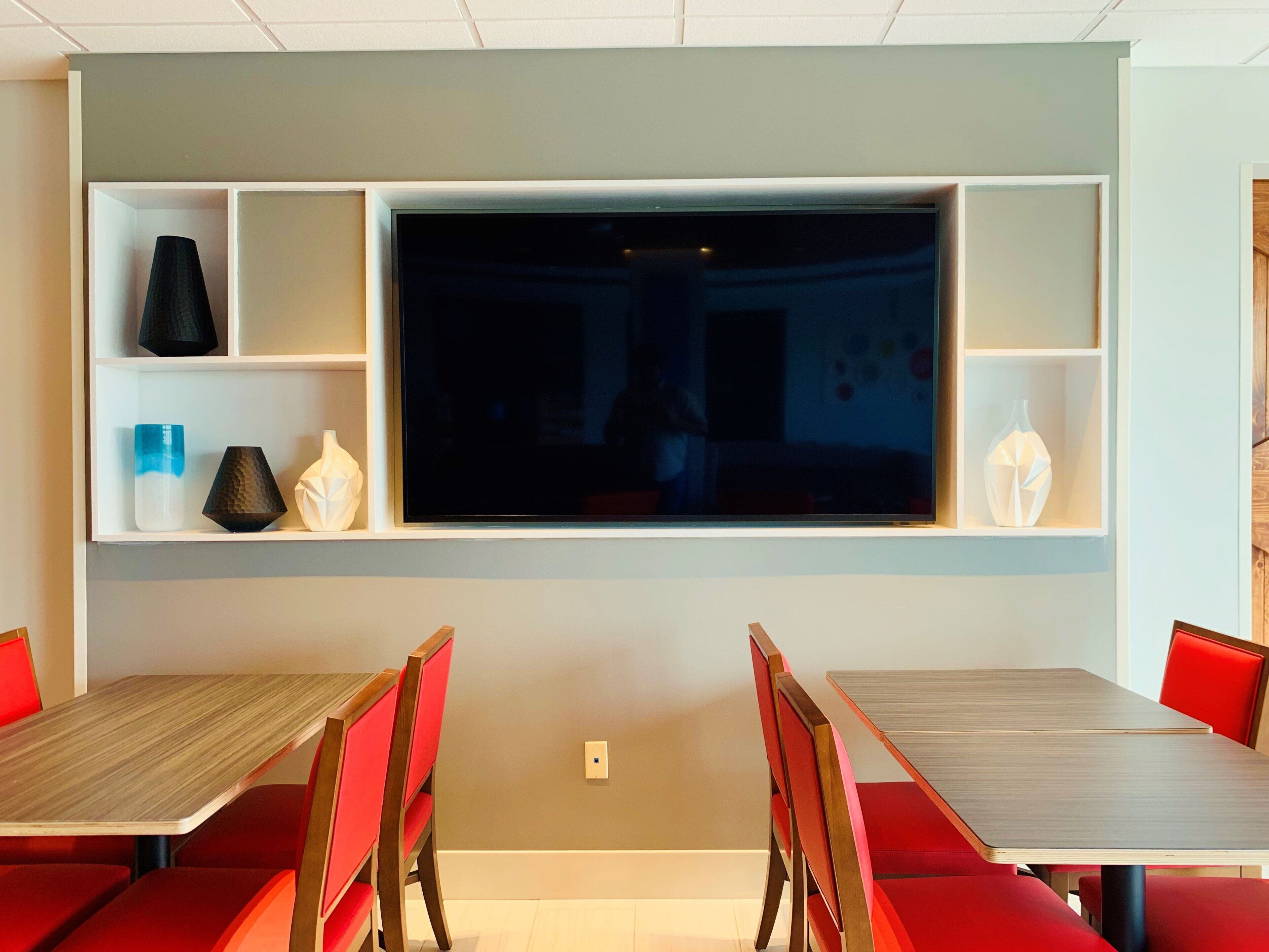 A dining area with a large flat-screen TV mounted on the wall, red upholstered chairs, and a wooden table.
