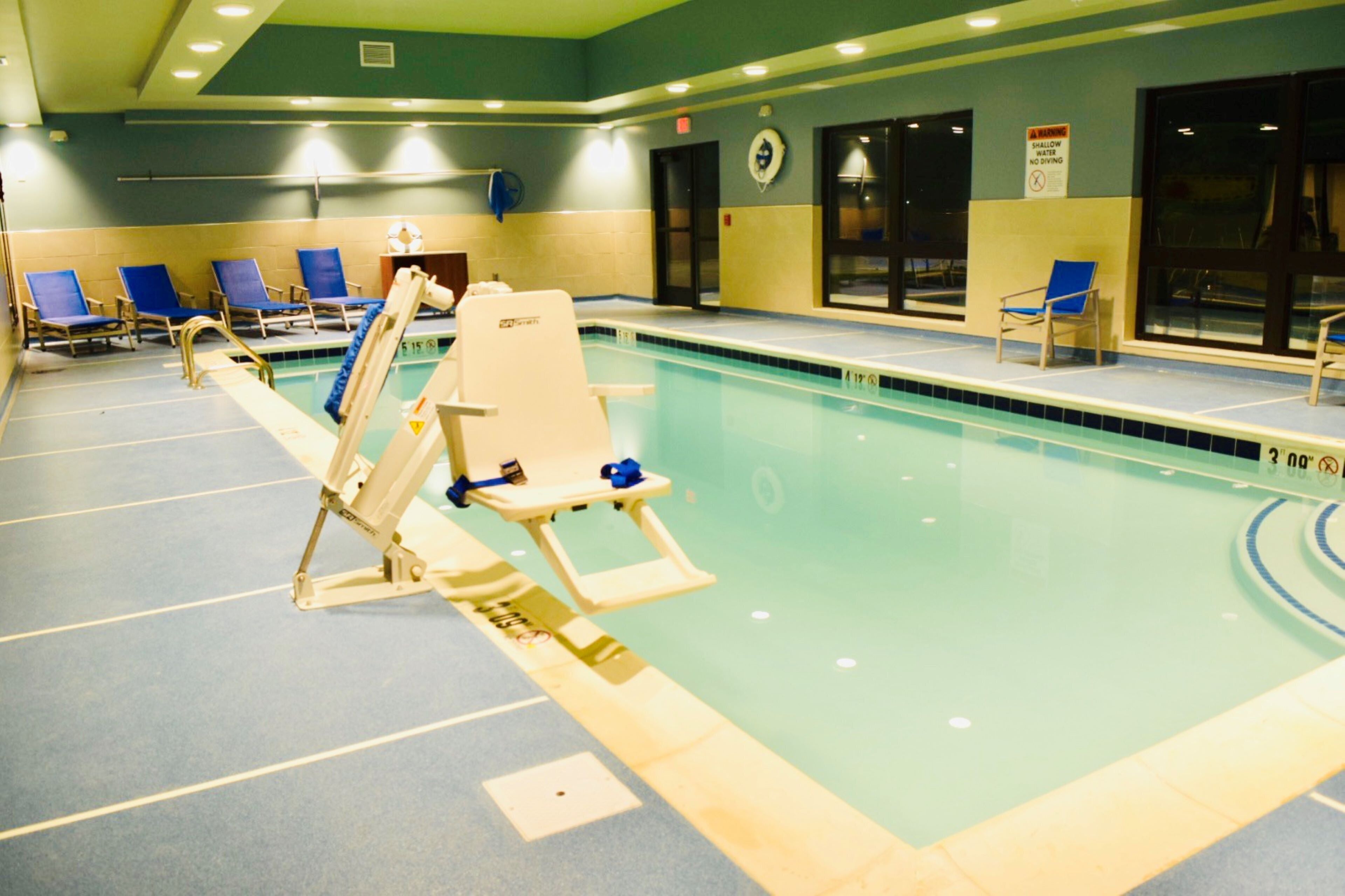 An indoor swimming pool with blue chairs, a lifeguard stand, and a shallow water warning sign.