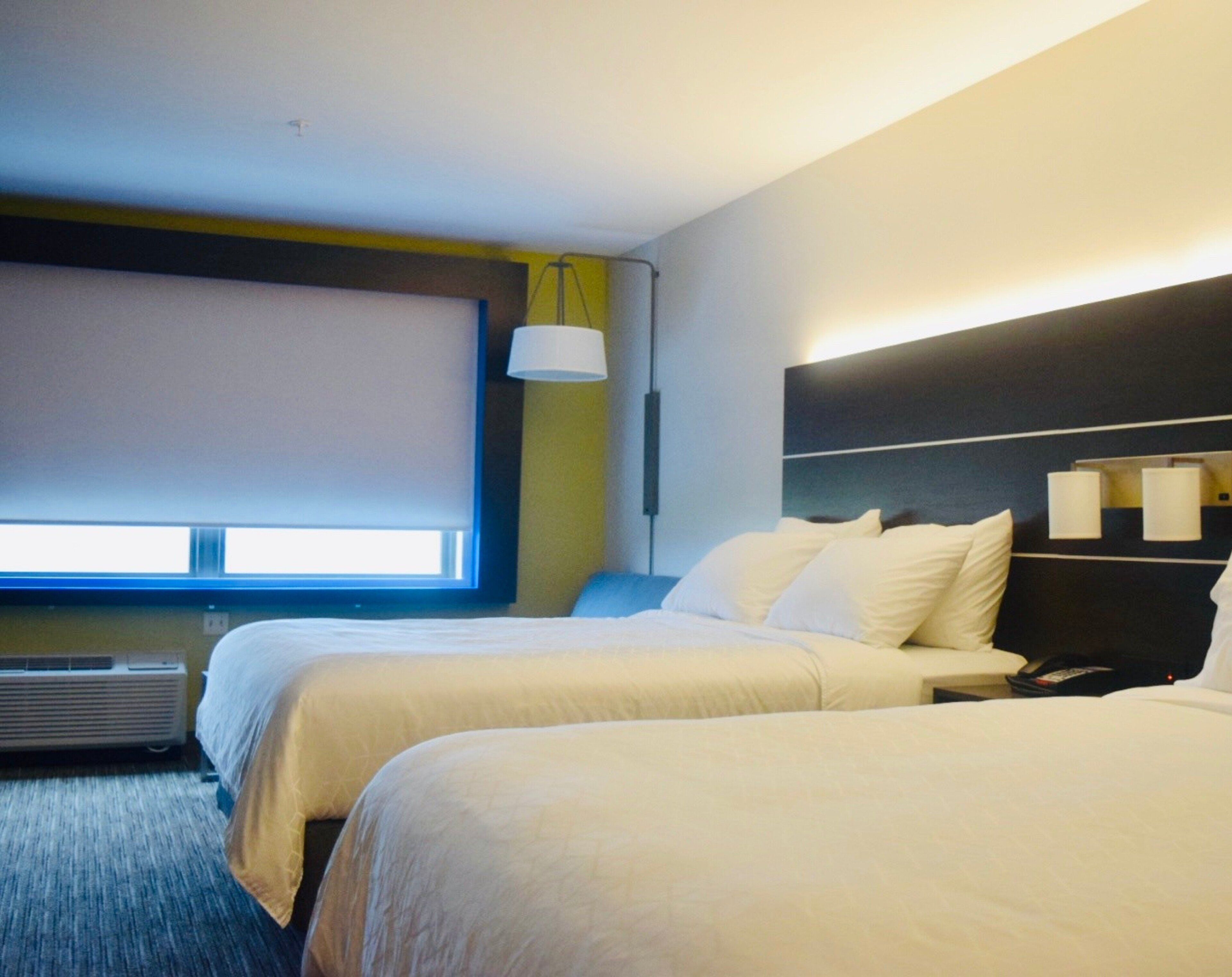A hotel room with two beds, a large window, an air conditioning unit, and a wall-mounted lamp.