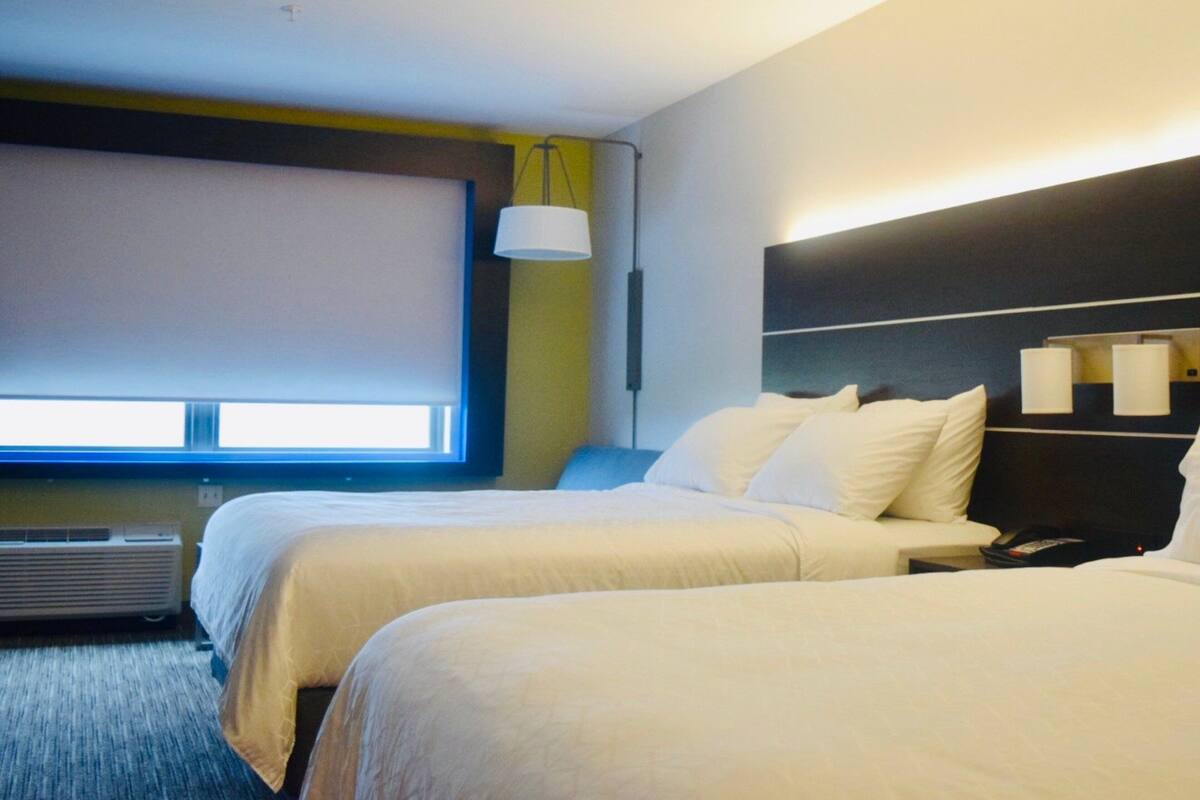 A hotel room with two beds, a large window, a wall-mounted lamp, and a ceiling light.