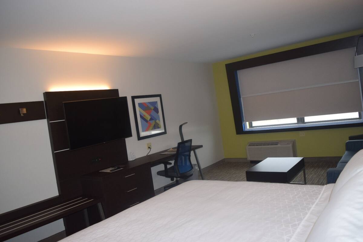 A hotel room with a bed, a desk, a chair, a TV, a window with blinds, and a picture on the wall.