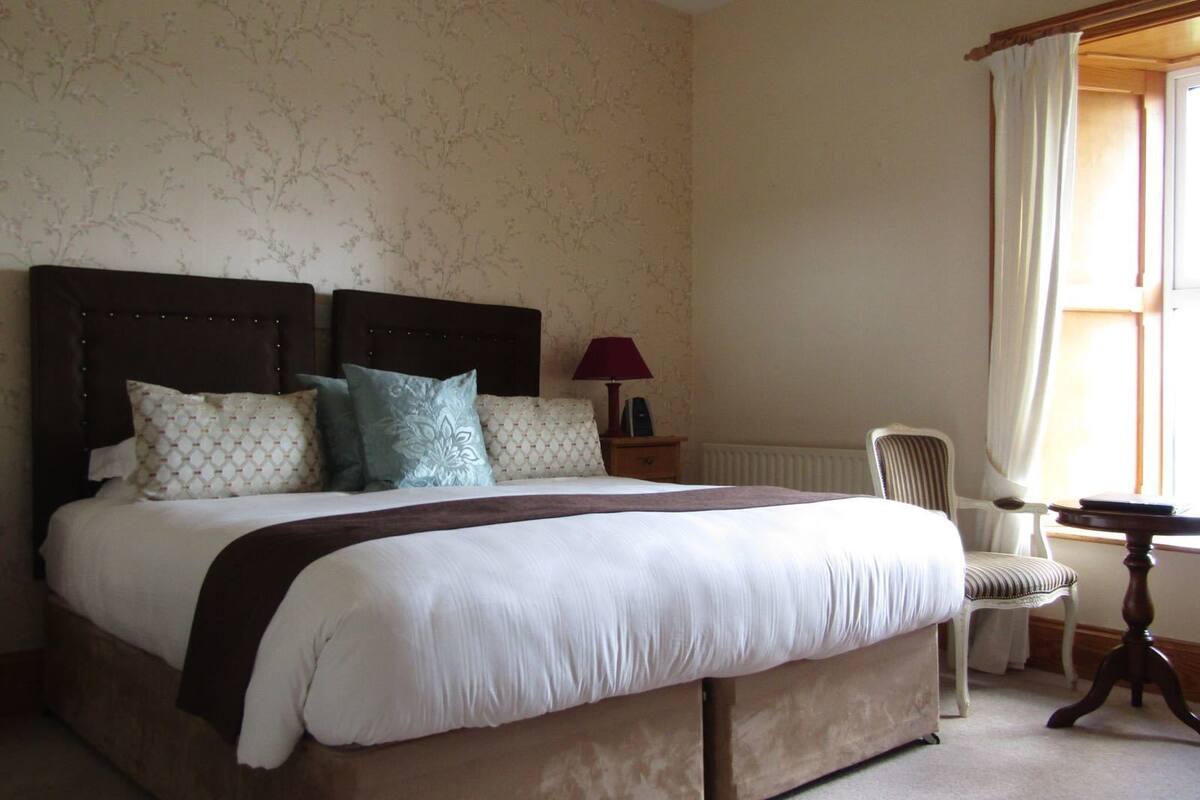 A neatly made bed with a dark headboard, a bedside table with a lamp, a chair, and a window with curtains.