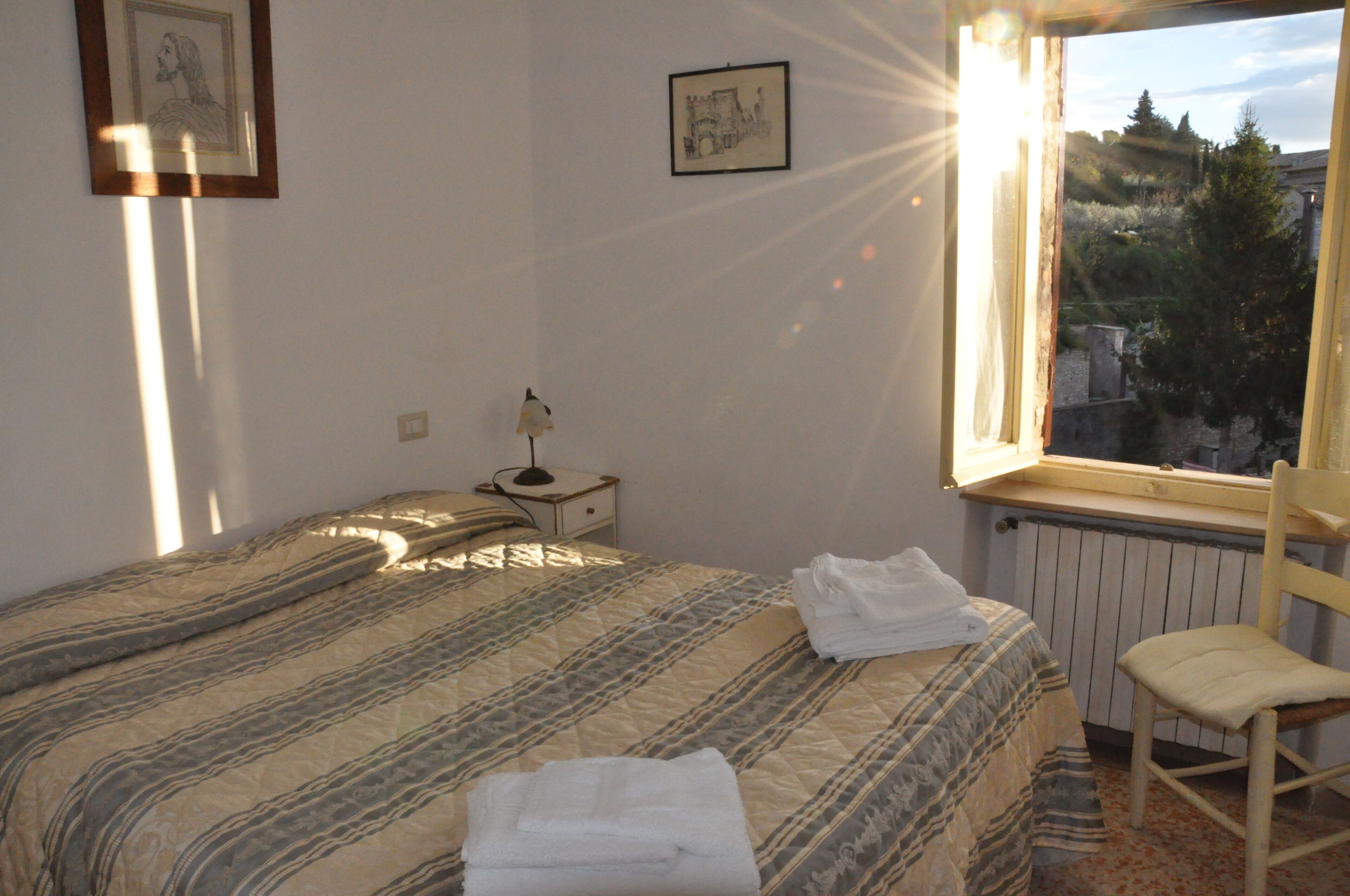 A bedroom with a bed, a chair, a nightstand, and a window with a view of trees and buildings.