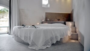 Un dormitorio moderno con una cama grande, un banco empotrado, un ventanal pequeño y un baño con espejo y lavamanos.