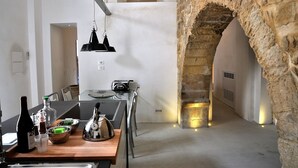 Una cocina estrecha con un arco de piedra, una lámpara colgante y una mesa de madera con utensilios de cocina.