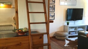 Eine Holzleiter lehnt an einer Wand in einem Raum mit Holzboden, einer Küchenzeile und einem an der Wand befestigten Fernseher.