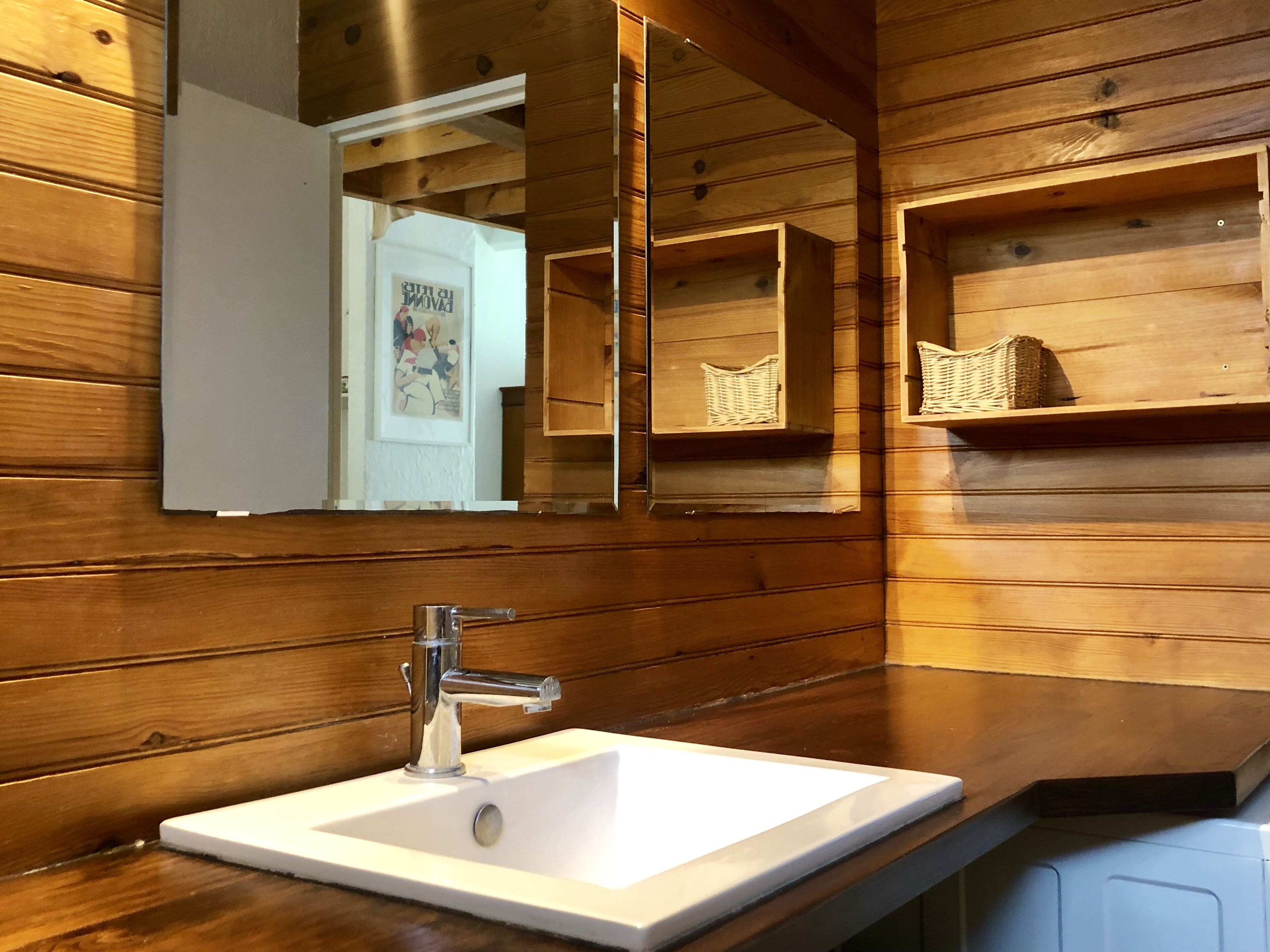 Une salle de bain avec un lambris en bois, un lavabo blanc doté d’un robinet chromé et un miroir qui reflète la pièce.