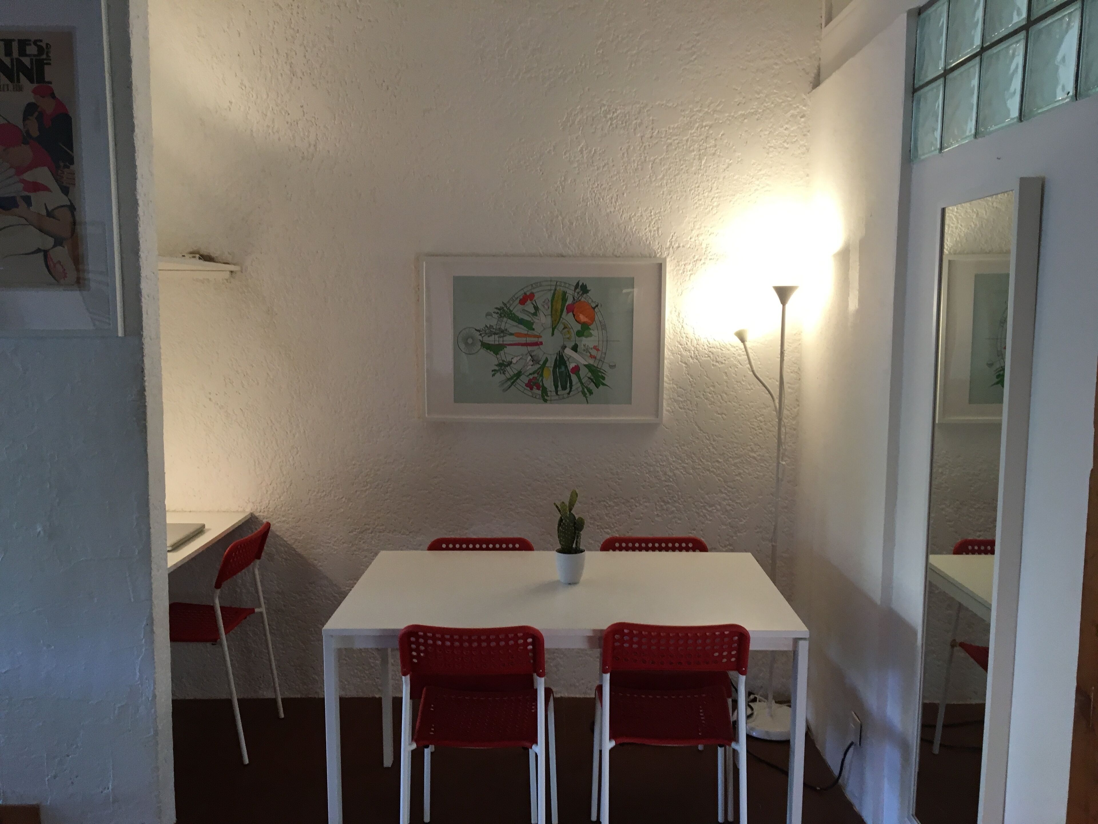 A dining area with a white table and red chairs, a framed artwork on the wall, and a tall floor lamp.