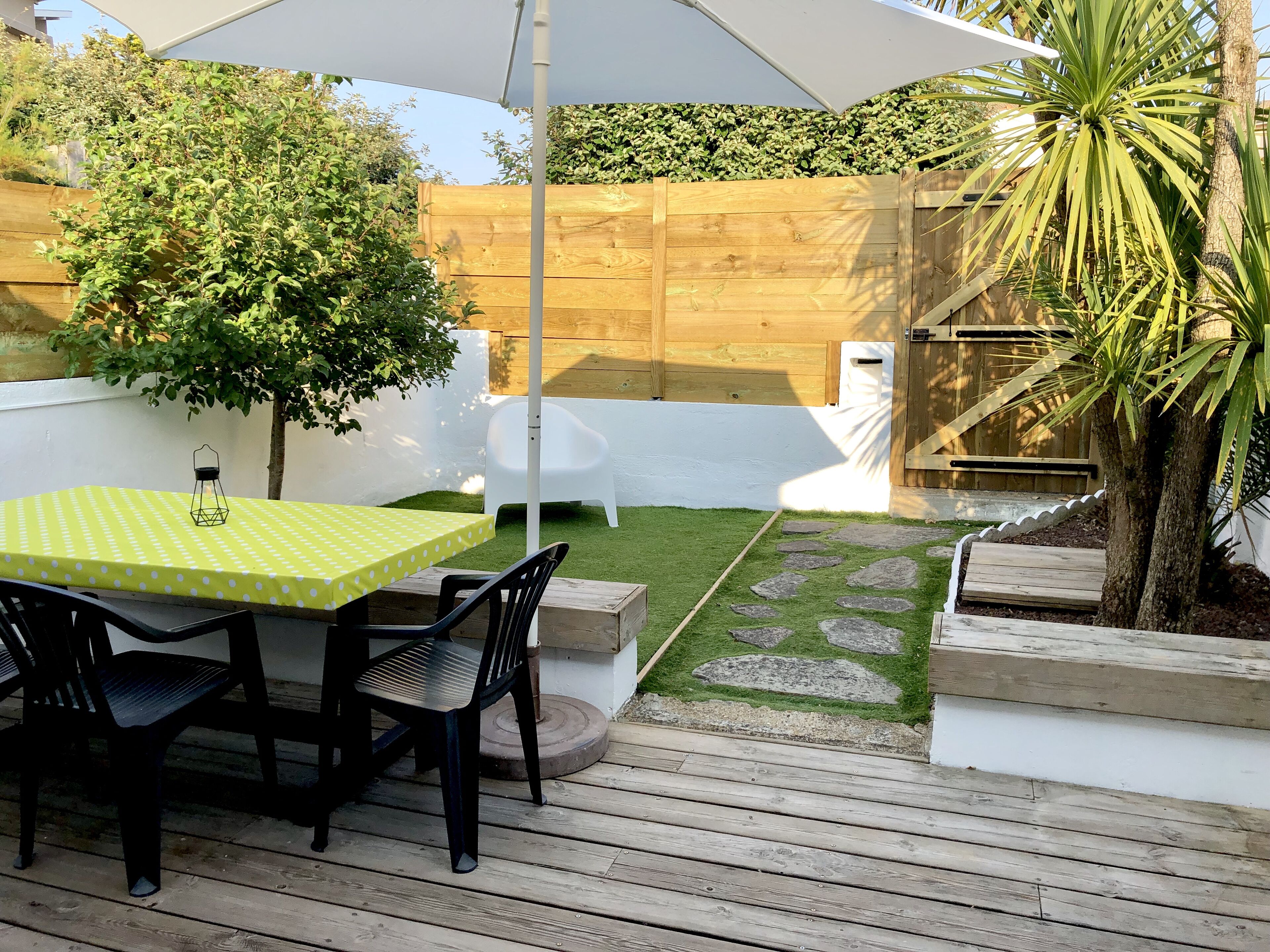 A patio area with a table and chairs, a wooden deck, a white fence, and a small garden with a palm tree.