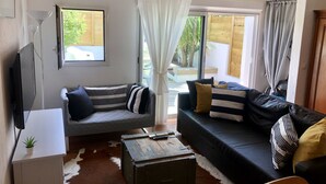 Ein modernes Wohnzimmer mit einer Couch, einem Couchtisch und einem Fernseher. Es gibt Fenster mit Vorhängen, einen Ausblick ins Freie und eine Deckenleuchte.
