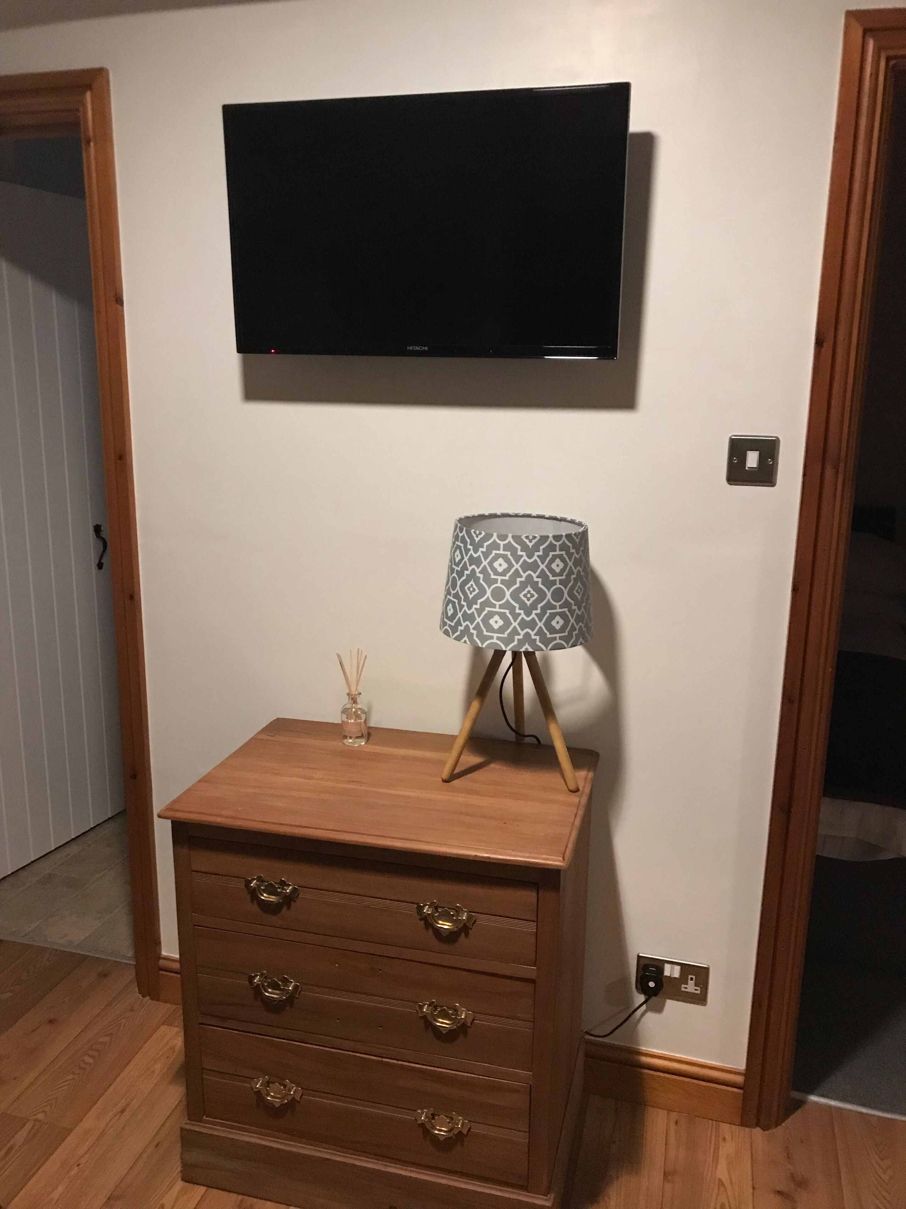 A wooden dresser with drawers and a lamp on top, a flat-screen TV mounted above it, and a power outlet on the wall.