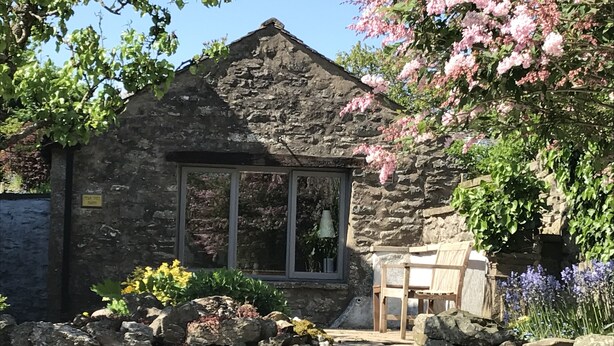A stone cottage with a large window, a stone staircase leading to a wooden door, and a garden with various plants and flowers. - Traditional Lakeland Hideaway Cottage for two, close to the Lakes and Dales (KENDAL)