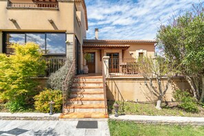 Una casa de dos pisos con una escalera que conduce a la entrada, un patio con muebles de exterior y un jardín con diversas plantas.