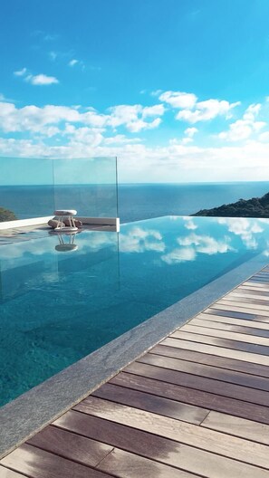 Una piscina infinita de color azul cristalino con una terraza de madera, con vistas a un paisaje montañoso y al mar.