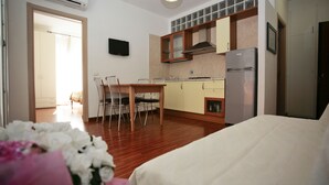 A compact hotel room with a bed, a dining table with chairs, a kitchen area with a refrigerator, and a TV mounted on the wall.