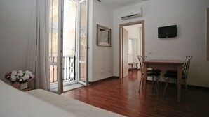 A hotel room with a wooden floor, a bed, a dining table with chairs, a TV mounted on the wall, and a door leading to a balcony.