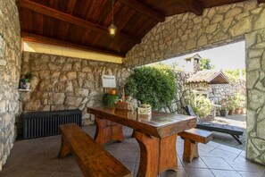 A stone-walled outdoor seating area with wooden benches and a table, a stone fireplace, and a sign reading "Dobrodosli".