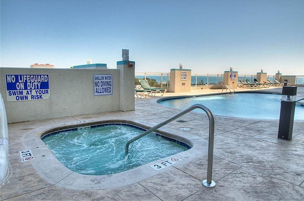 A rooftop pool area with a hot tub, lounge chairs, and warning signs about no lifeguard and shallow water.