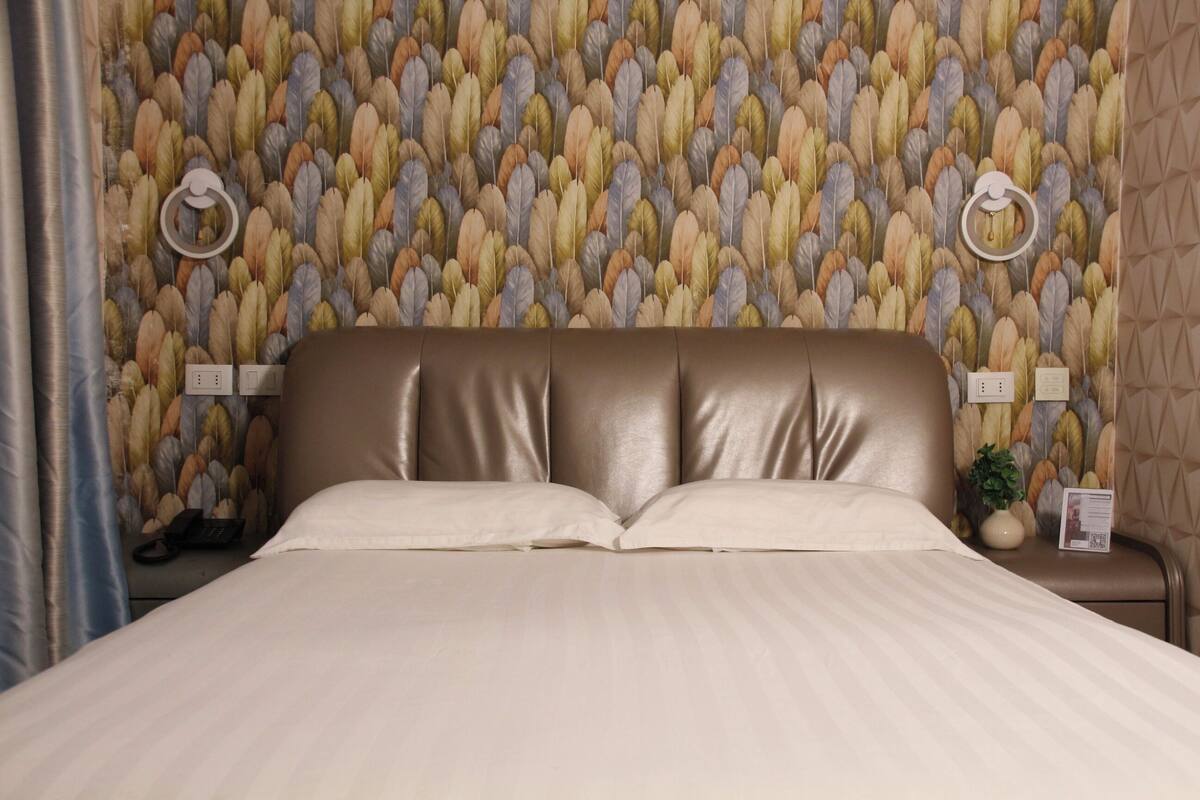 Quarto de hotel com cama, cabeceira, parede com padrão de penas, duas mesas de cabeceira com luminárias e uma cortina.