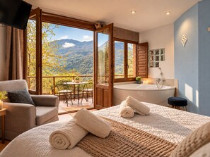A bedroom with a large bed, a sitting area with a table and chairs, a bathtub, and a balcony with a mountain view. - Magical Place, Ideal Couples: Jacuzzi, Mountains, Barbecue And Fireplace (Asturias)