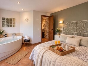 A bedroom with a large bed, a bedside table with a lamp, a nightstand, a wall-mounted clock, and a door leading to a bathroom with a bathtub. - Magical Place, Ideal Couples: Jacuzzi, Mountains, Barbecue And Fireplace (Asturias)