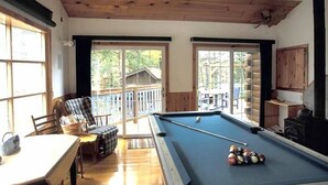 Une salle de billard équipée d’une table de billard, de chaises et offrant une vue sur une terrasse et des arbres.