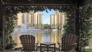A gazebo with two Adirondack chairs and a small table, overlooking a fountain and a body of water with high-rise buildings in the background.