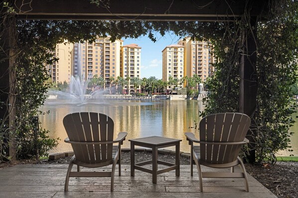 A gazebo with two Adirondack chairs and a small table, overlooking a fountain and a body of water with high-rise buildings in the background.