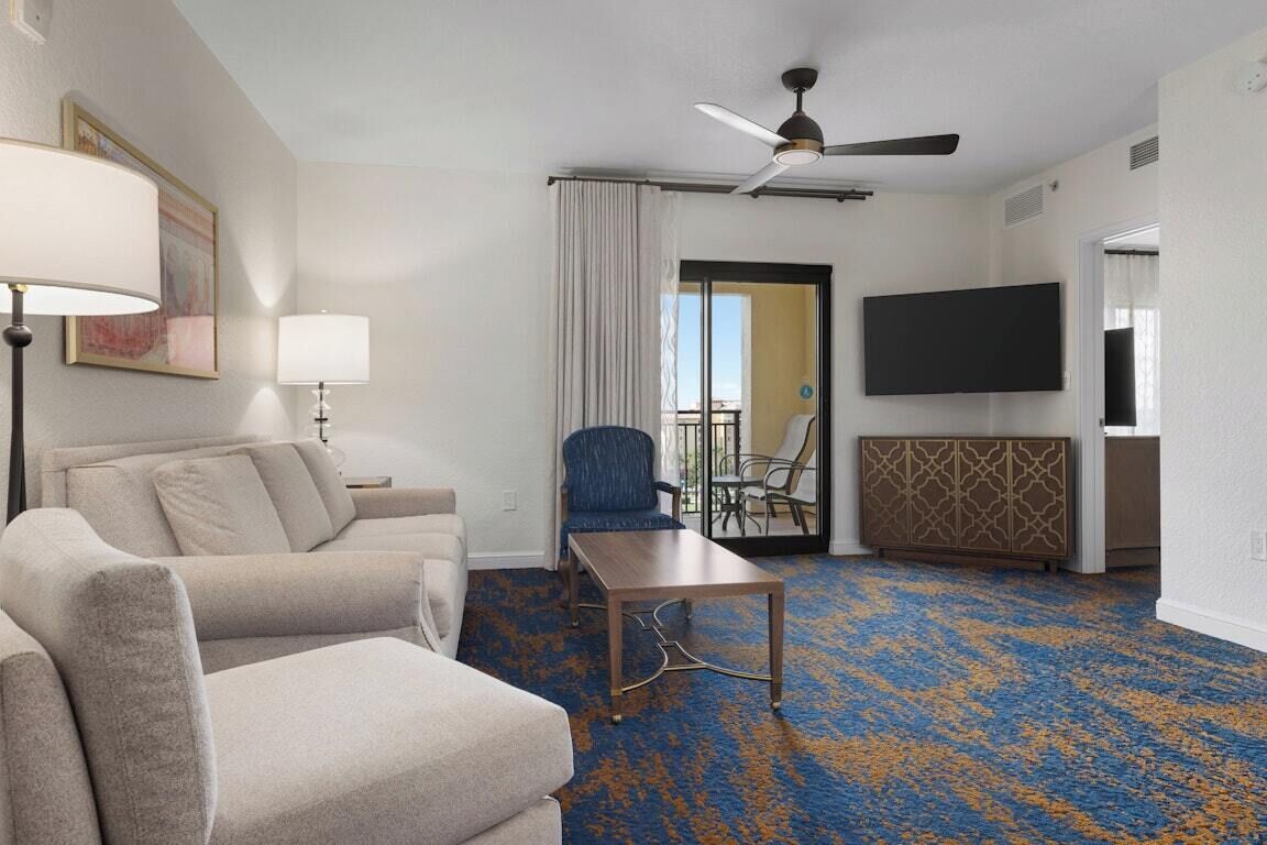A hotel room with a sofa, a coffee table, a ceiling fan, a TV, and a patterned carpet.