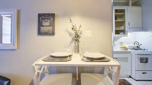 Un coin repas avec une table blanche dressée pour deux personnes, un vase rempli de fleurs et, au fond, une cuisine avec des meubles et des appareils électroménagers blancs.