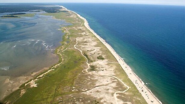 Vista aérea de una zona costera con playa, carretera y cuerpo de agua.