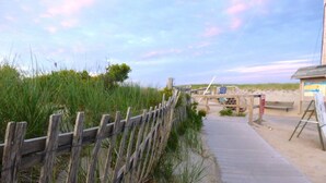 Un paseo marítimo de madera que lleva a una zona de playa, con una valla de madera, un banco y un caballete con una pintura.