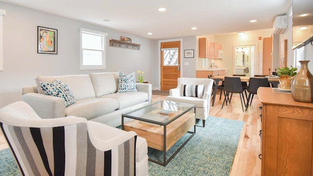 A modern living room with a sofa, armchairs, and a coffee table. There is a dining area with a table and chairs, and a kitchen in the background.
