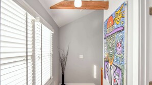 A hallway with white blinds, a wooden floor, a black vase, and a colorful wall mural.