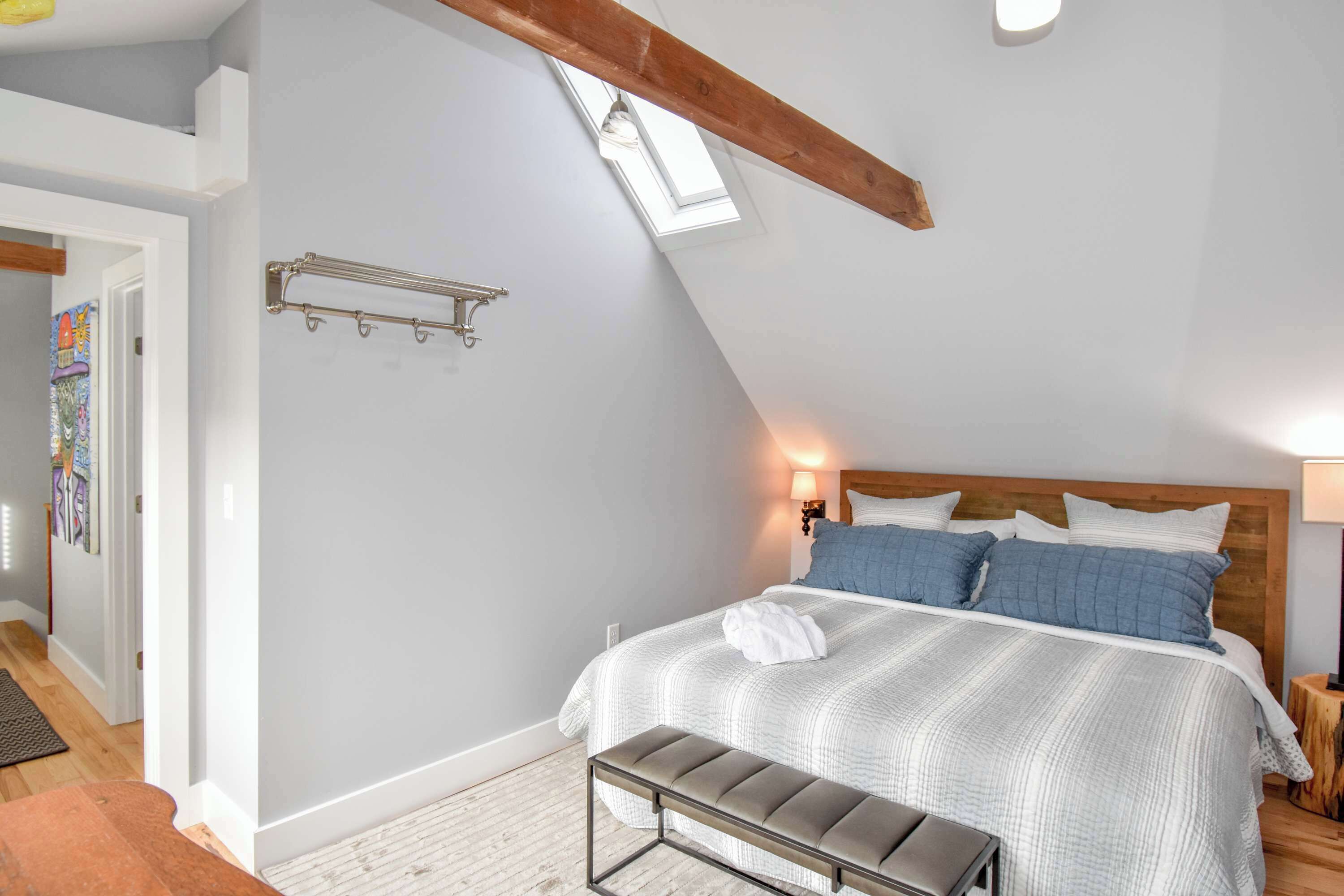 Une chambre avec un plafond en pente, une tête de lit en bois, un lit avec une literie blanche et bleue, un banc et un puits de lumière.