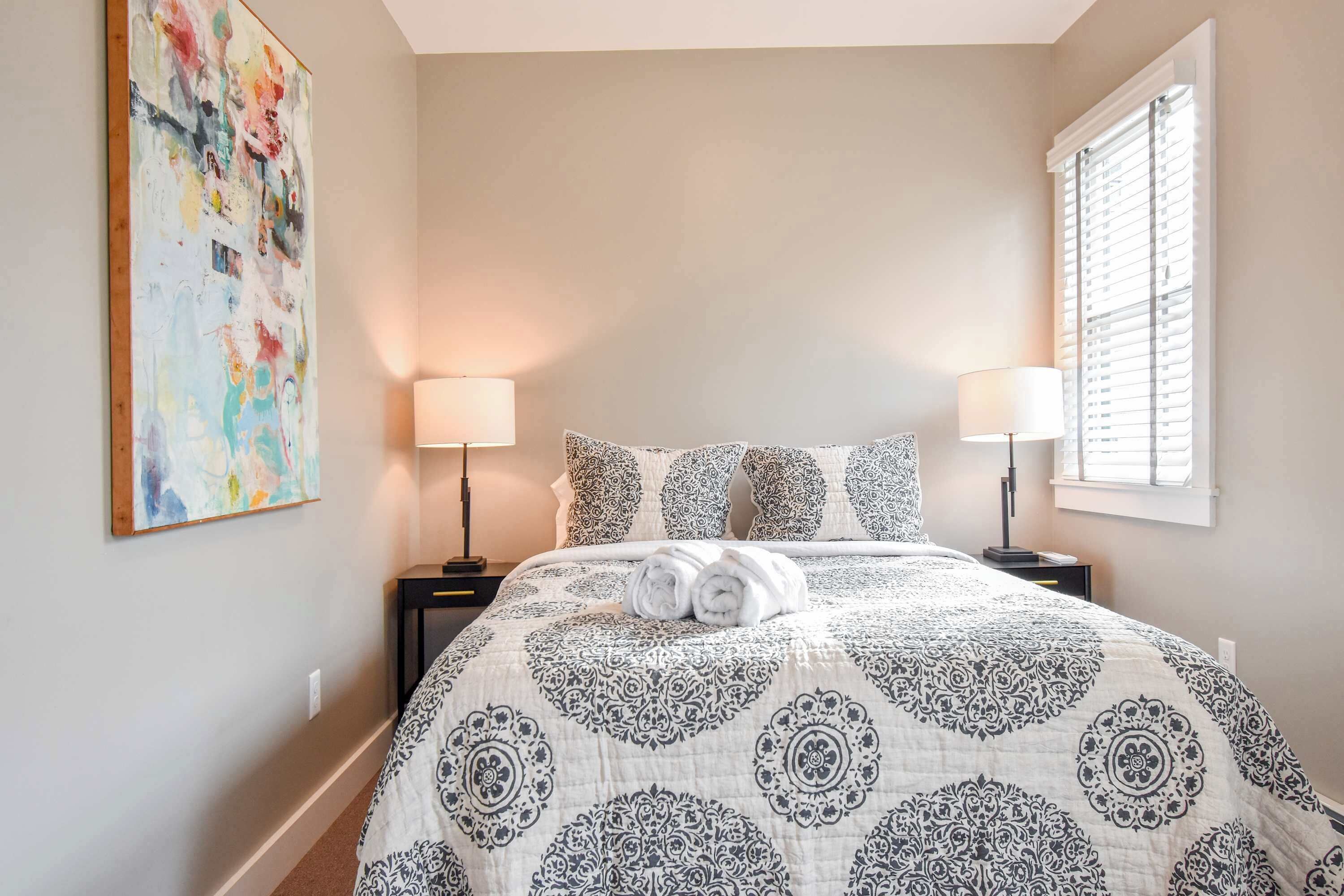 A neatly made bed with a patterned bedspread, two bedside lamps, and a large abstract painting on the wall.