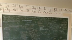 Et klasseværelse med en stor, grøn tavle fyldt med skrift og en whiteboard over den, der viser alfabetet.