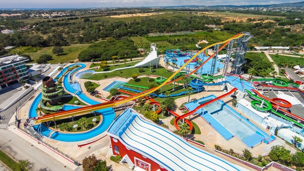 Parque acuático con múltiples toboganes y piscinas, rodeado de vegetación y un horizonte urbano a lo lejos.