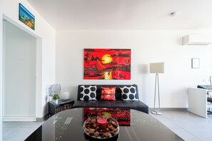 Un salon moderne avec une table basse en verre, un canapé agrémenté de coussins à motifs, un tableau représentant un coucher de soleil et un lampadaire.