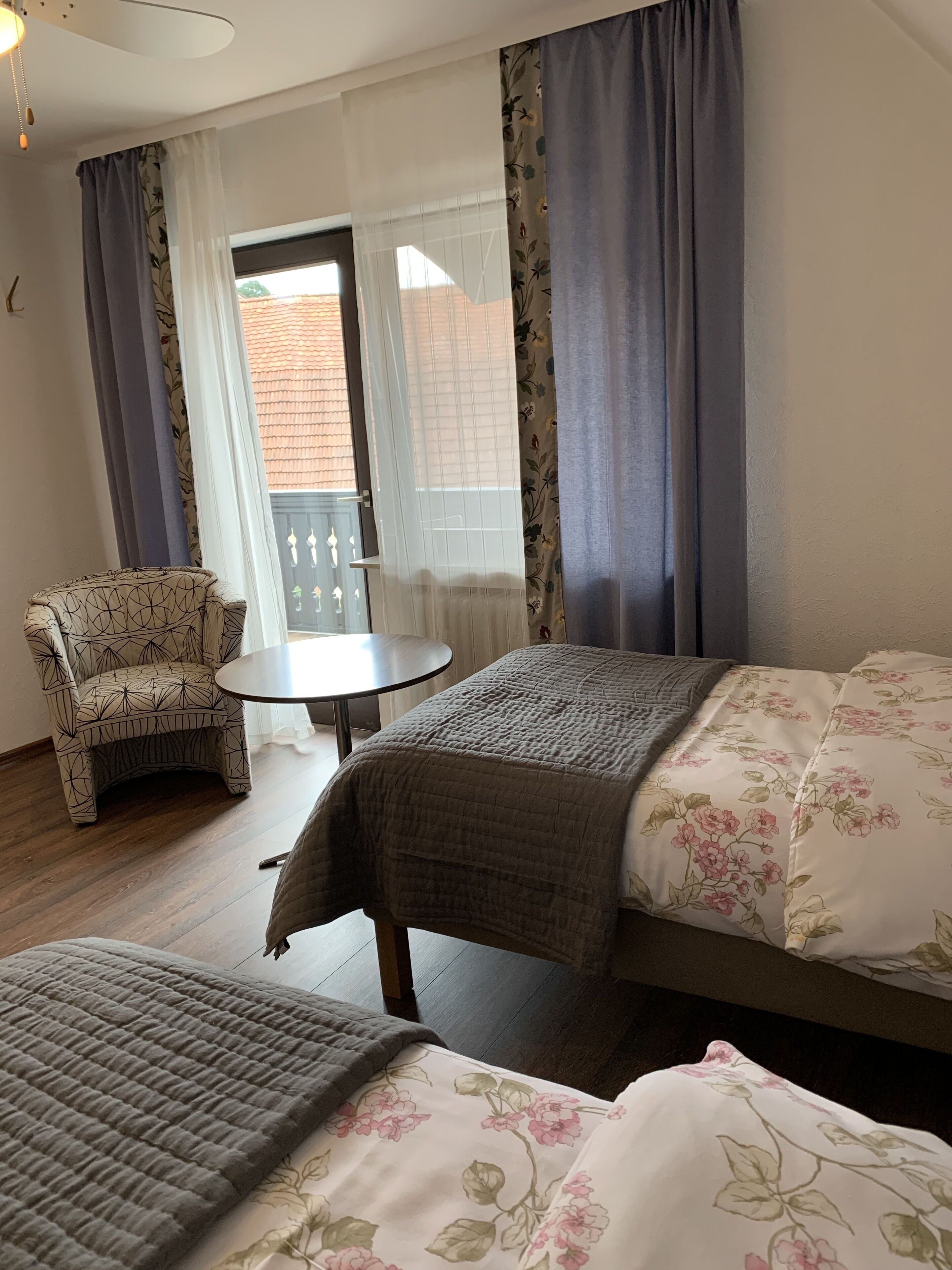 Ein Hotelzimmer mit zwei Betten, einem Sessel, einem kleinen Tisch und einem Balkon mit Blick auf ein Gebäude.