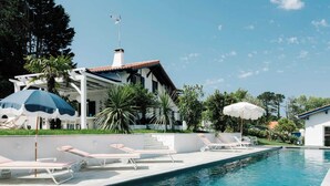 A swimming pool with lounge chairs and umbrellas, surrounded by greenery and a building with a tiled roof. - House center Guethary Maisons Dominos 1 (Guéthary)