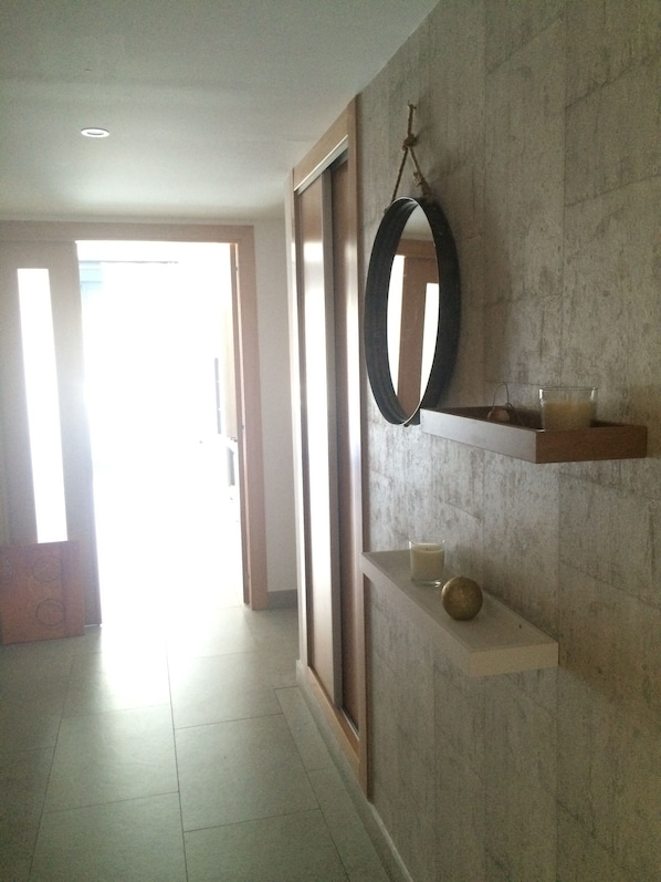 Un couloir avec un grand miroir, une petite étagère sur laquelle se trouve une bougie et un objet décoratif, et une porte qui donne sur une autre pièce.