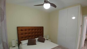 Een slaapkamer met een gestapeld hoofdbord, een bed met wit beddengoed en bruine kussens, een nachtkastje met een lamp, een plafondventilator en een garderobe.